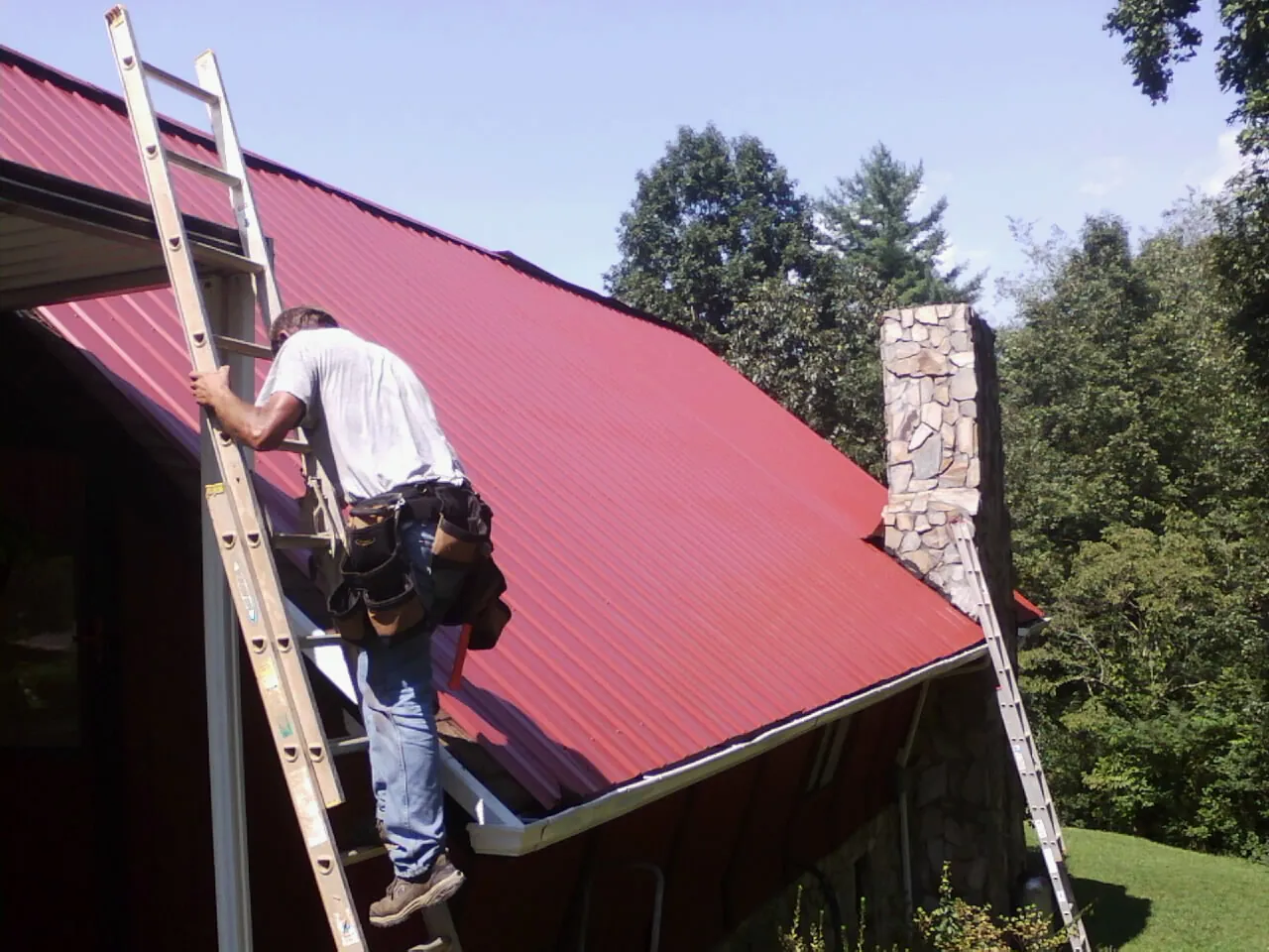 Roofer on a ladder beside a red metal roof and stone chimney — Soffit & Fascia Repair by Roofing Brothers in Mcclellan